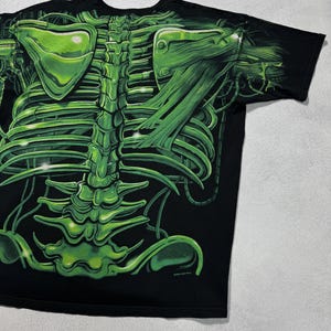 Puede incluir: Camiseta negra con un dise&ntilde;o de esqueleto verde vibrante. El gr&aacute;fico incluye detalles de la caja tor&aacute;cica, la columna vertebral y los om&oacute;platos, creando un efecto visual impactante. El dise&ntilde;o est&aacute; impreso sobre un fondo negro.