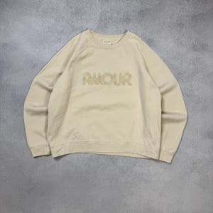 May include: A cream-colored long-sleeve sweatshirt featuring a crew neckline and raglan sleeves. The word AMOUR is embroidered in a matching cream tone across the center of the chest. The garment has ribbed cuffs and a ribbed hemline.