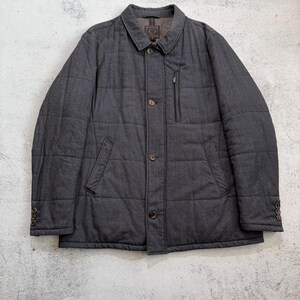 May include: A charcoal gray quilted jacket with a button-up front, a small chest pocket, and two side pockets. The jacket has a collared neckline and buttoned cuffs. The jacket is made of a textured fabric.