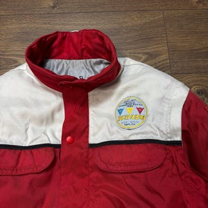 May include: Red and white jacket with a round patch that says "World Champion 500 CC 1986". The jacket has two front pockets and a red collar. The jacket has the word "Dainese" on the patch.