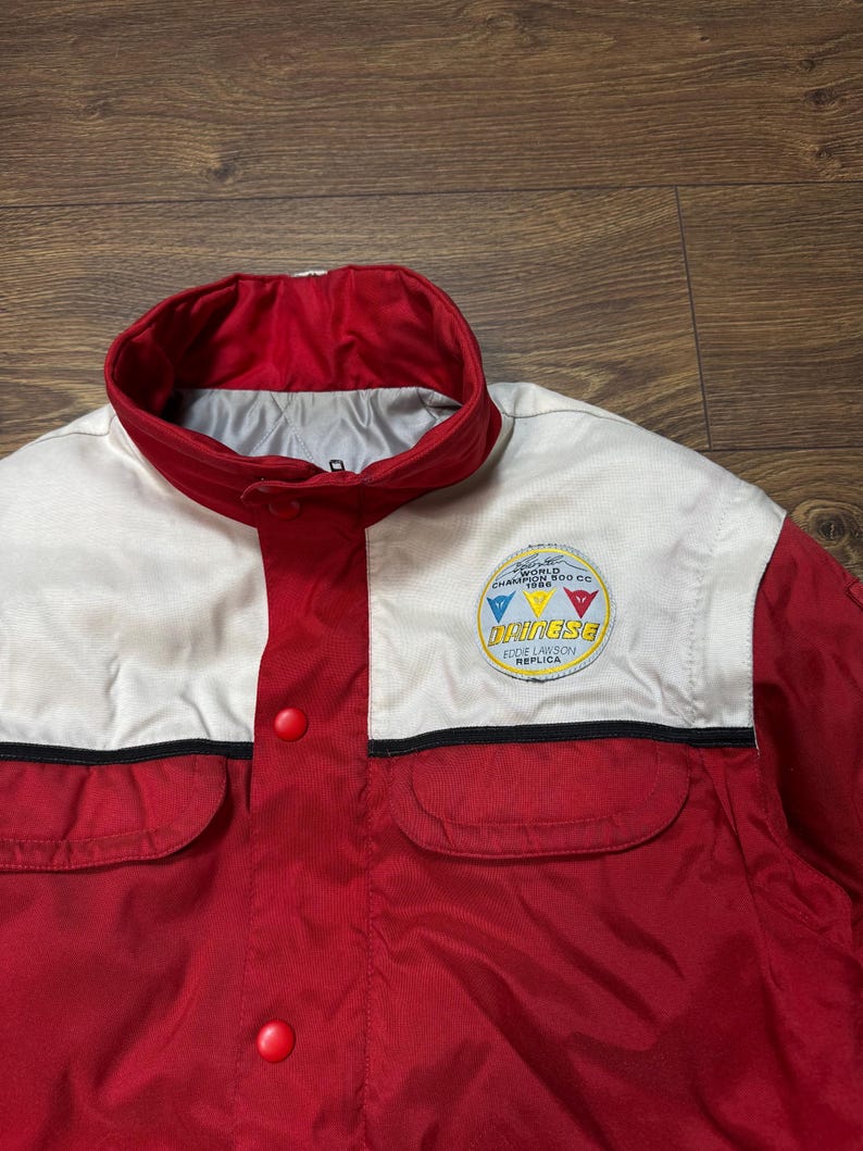 May include: Red and white jacket with a round patch that says "World Champion 500 CC 1986". The jacket has two front pockets and a red collar. The jacket has the word "Dainese" on the patch.