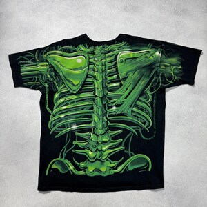 Puede incluir: Camiseta negra con un dise&ntilde;o esquel&eacute;tico verde ne&oacute;n. El gr&aacute;fico incluye una representaci&oacute;n detallada de una caja tor&aacute;cica, columna vertebral y otros elementos anat&oacute;micos, con una est&eacute;tica futurista y mec&aacute;nica. El dise&ntilde;o est&aacute; impreso sobre un fondo negro.