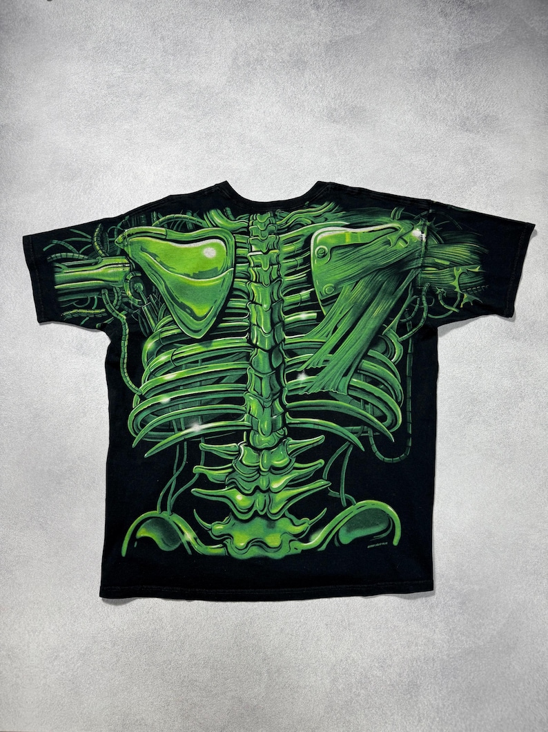 Puede incluir: Camiseta negra con un dise&ntilde;o esquel&eacute;tico verde ne&oacute;n. El gr&aacute;fico incluye una representaci&oacute;n detallada de una caja tor&aacute;cica, columna vertebral y otros elementos anat&oacute;micos, con una est&eacute;tica futurista y mec&aacute;nica. El dise&ntilde;o est&aacute; impreso sobre un fondo negro.