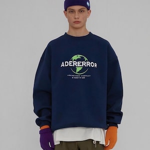 May include: Navy blue sweatshirt with the text "ADERERROR" above a green globe graphic. The sweatshirt also has the text "ADERERROR COMPANY" below the globe. The person is wearing a purple and orange gloves and a purple and green beanie.