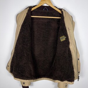 May include: Tan jacket with a brown, fuzzy interior, hanging on a wooden hanger. The jacket has a zipper closure, button details, and a small label with the brand name. The jacket is a neutral color and appears to be a warm, outerwear garment.