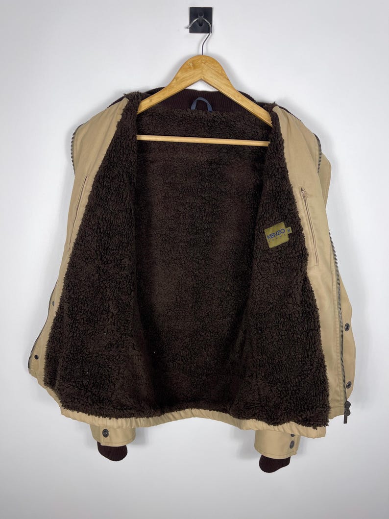 May include: Tan jacket with a brown, fuzzy interior, hanging on a wooden hanger. The jacket has a zipper closure, button details, and a small label with the brand name. The jacket is a neutral color and appears to be a warm, outerwear garment.