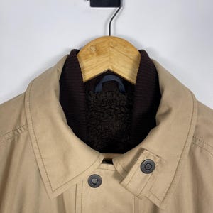 May include: A tan jacket with a dark brown collar and lining, hanging on a wooden hanger. The jacket has two dark buttons and a classic design, suitable for outerwear.
