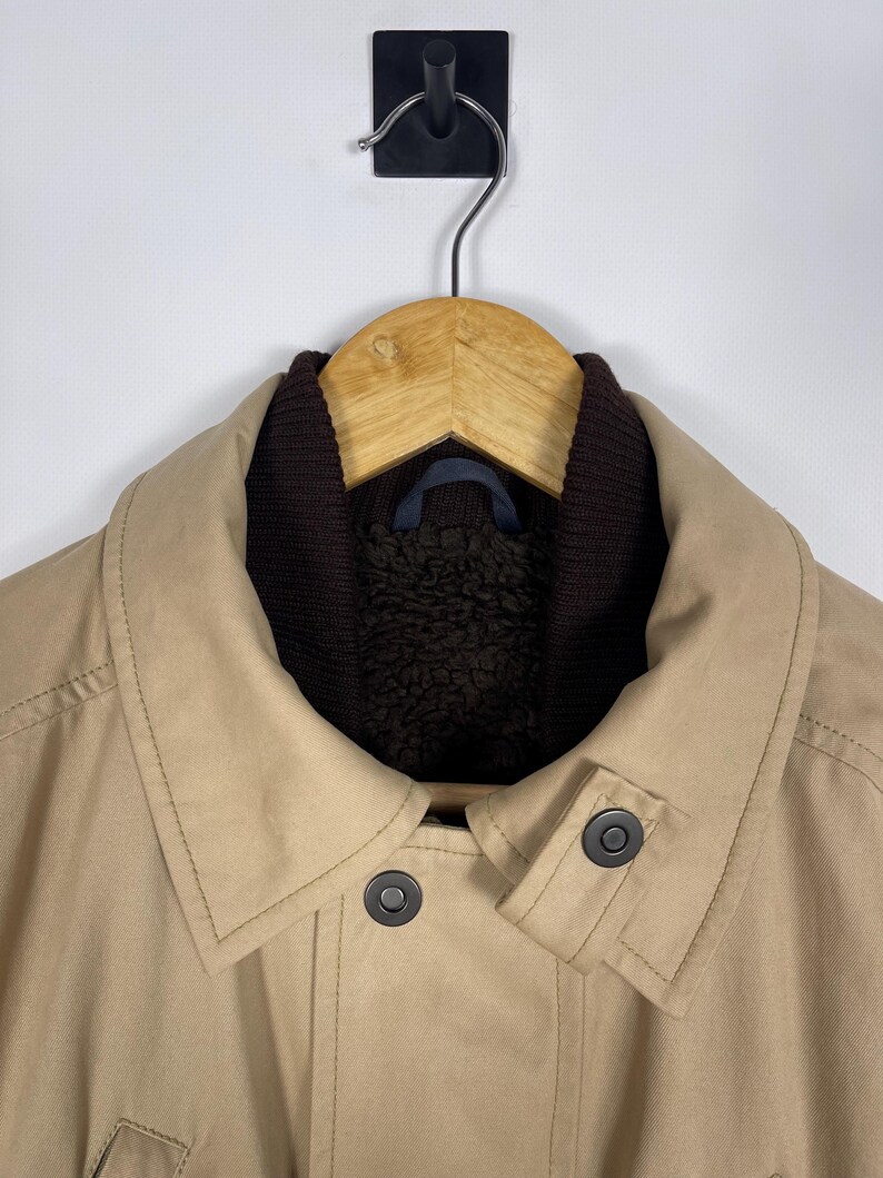 May include: A tan jacket with a dark brown collar and lining, hanging on a wooden hanger. The jacket has two dark buttons and a classic design, suitable for outerwear.
