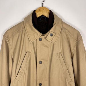 May include: A tan-colored coat with a brown collar and a button-up front. The coat has two front pockets and is hanging on a wooden hanger. The coat appears to be made of a durable material, suitable for cooler weather.