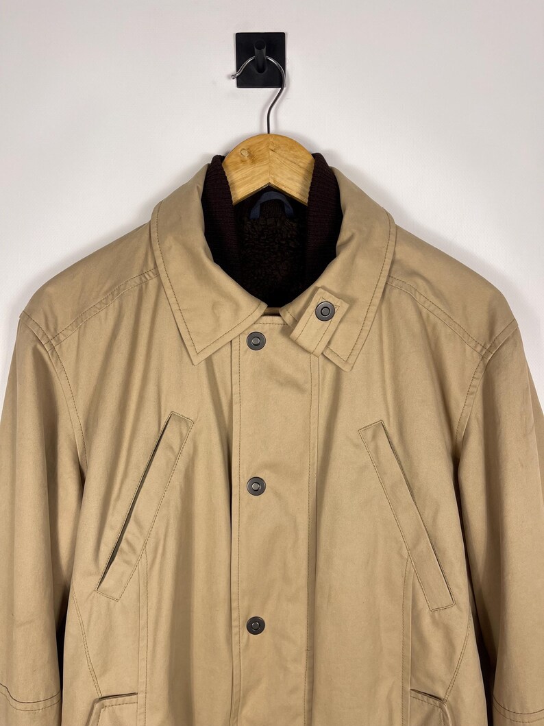 May include: A tan-colored coat with a brown collar and a button-up front. The coat has two front pockets and is hanging on a wooden hanger. The coat appears to be made of a durable material, suitable for cooler weather.