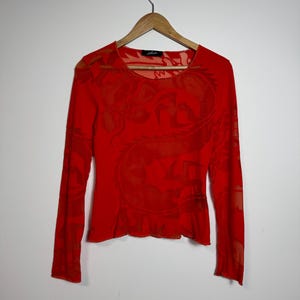 May include: Vibrant red long-sleeved top with a round neck and a delicate pattern. The sheer material reveals the hanger behind it. The top is displayed on a wooden hanger against a plain white backdrop.