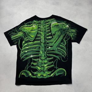 Puede incluir: Camiseta negra con un dise&ntilde;o de esqueleto verde vibrante. El gr&aacute;fico incluye una detallada caja tor&aacute;cica, columna vertebral y om&oacute;platos, creando un efecto visual impactante. La camiseta es de cuello redondo y manga corta.