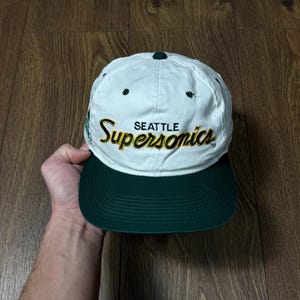 May include: A white and green Seattle SuperSonics baseball cap. The cap has the team name in gold lettering, a green peak, and a dark green button. This vintage cap is a classic style.