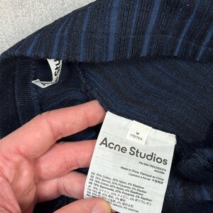 Puede incluir: Primer plano de una prenda de punto acanalado azul marino con una etiqueta de Acne Studios. La etiqueta incluye el nombre de la marca, la talla M y los detalles de la composici&oacute;n de la tela. La prenda est&aacute; hecha en China.