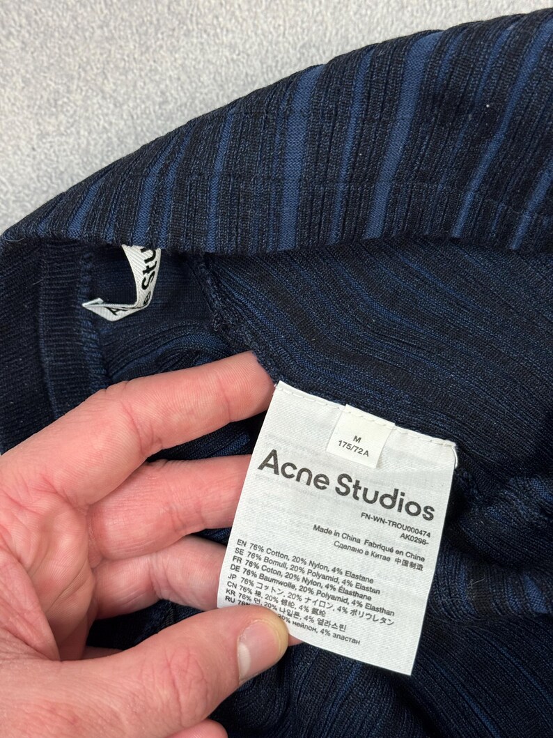Puede incluir: Primer plano de una prenda de punto acanalado azul marino con una etiqueta de Acne Studios. La etiqueta incluye el nombre de la marca, la talla M y los detalles de la composici&oacute;n de la tela. La prenda est&aacute; hecha en China.