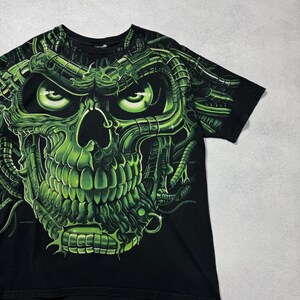 Puede incluir: Camiseta negra con un dise&ntilde;o de calavera verde ne&oacute;n. La calavera tiene una apariencia mec&aacute;nica detallada con ojos verdes brillantes y detalles intrincados. El dise&ntilde;o cubre la parte delantera de la camiseta, creando un efecto visual llamativo.