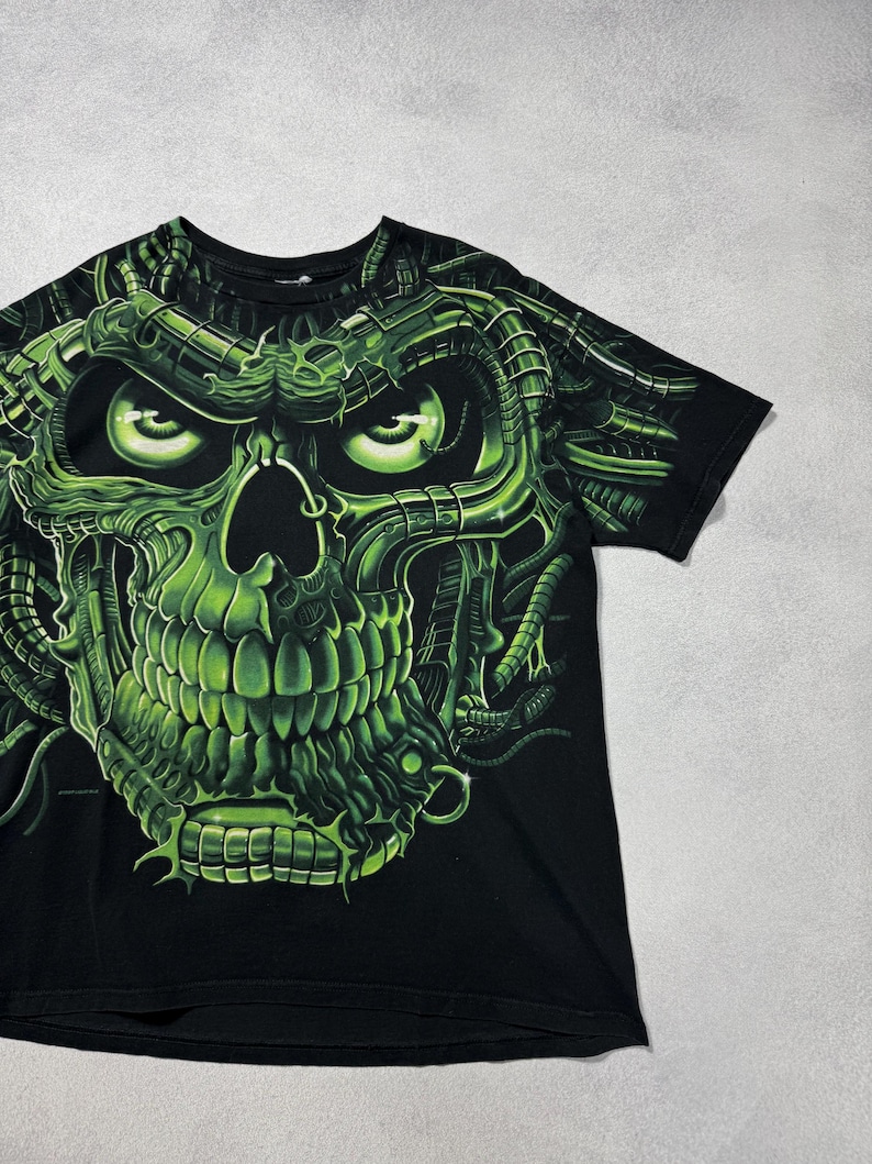 Puede incluir: Camiseta negra con un dise&ntilde;o de calavera verde ne&oacute;n. La calavera tiene una apariencia mec&aacute;nica detallada con ojos verdes brillantes y detalles intrincados. El dise&ntilde;o cubre la parte delantera de la camiseta, creando un efecto visual llamativo.
