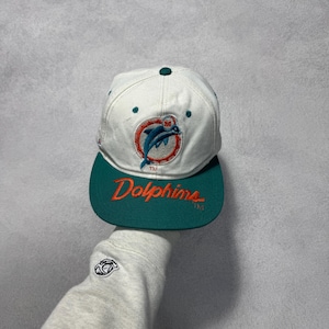 Op de afbeelding: Een witte en teal Dolphins baseball cap. De cap heeft het logo van het team op de voorkant, met het woord "Dolphins" in oranje letters op de klep. De cap wordt door een persoon omhoog gehouden.
