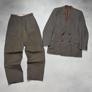 以下が含まれることがあります： 茶色のピンストライプのスーツ。ダブルブレストのブレザーと、お揃いのパンツが含まれています。ブレザーは濃い色のボタンと茶色の裏地付き。パンツはプリーツフロントです。スーツは明るい灰色の表面に置かれています。
