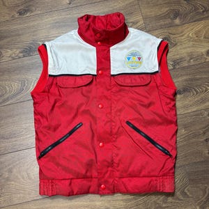 May include: A red and white vintage vest with a high collar and two front pockets. The vest has a white upper section with a logo and red accents. Features black zippers and red buttons.