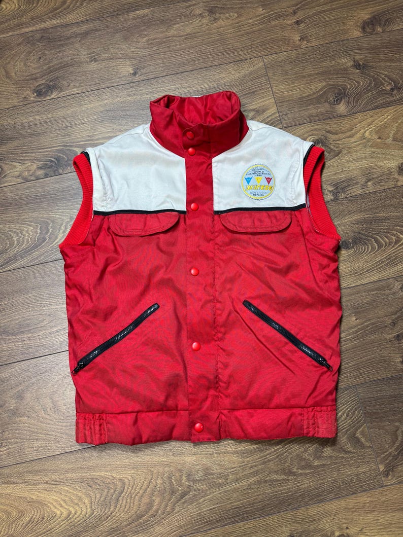 May include: A red and white vintage vest with a high collar and two front pockets. The vest has a white upper section with a logo and red accents. Features black zippers and red buttons.