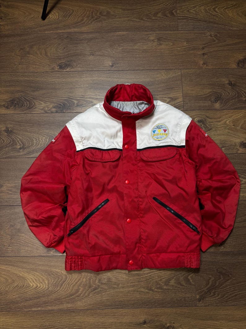 May include: Red and white vintage jacket with a stand-up collar and a small logo on the chest. The jacket has a button-up front, two front pockets with black zippers, and a ribbed hem and cuffs. The jacket is made of a shiny material.