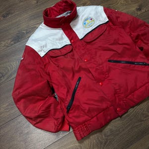 May include: Red and white vintage jacket with a stand-up collar, two front pockets, and a zipper closure. The jacket has a logo patch on the chest and a ribbed hem and cuffs. The jacket is made of a lightweight material.