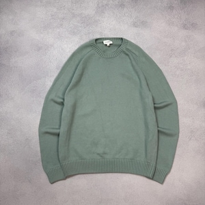 May include: A sage green long-sleeve sweater featuring a classic crew neckline, ribbed cuffs, and a matching ribbed hem. The garment is constructed with a fine knit texture and includes a small white brand label stitched inside the collar.