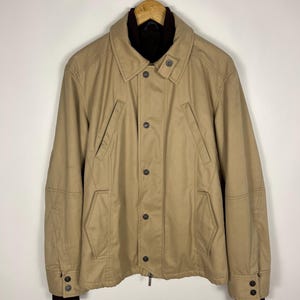 May include: Tan jacket with a dark brown collar and cuffs. The jacket has a button-up front, two front pockets, and a zipper at the bottom. The jacket is hanging on a wooden hanger.