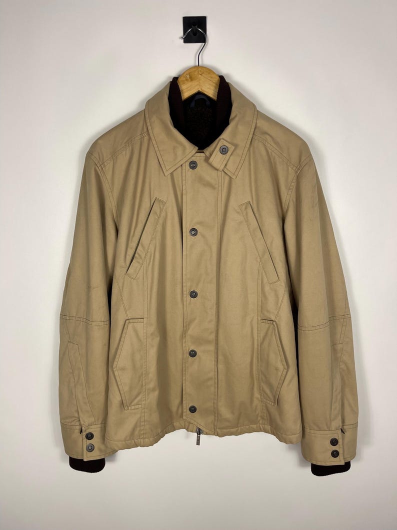 May include: Tan jacket with a dark brown collar and cuffs. The jacket has a button-up front, two front pockets, and a zipper at the bottom. The jacket is hanging on a wooden hanger.