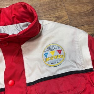 May include: Red and white racing jacket with a button closure and a small pocket. The jacket features a patch with the text "Dainese Eddie Lawson Replica" and a logo with the text "World Champion 500 CC 1986".