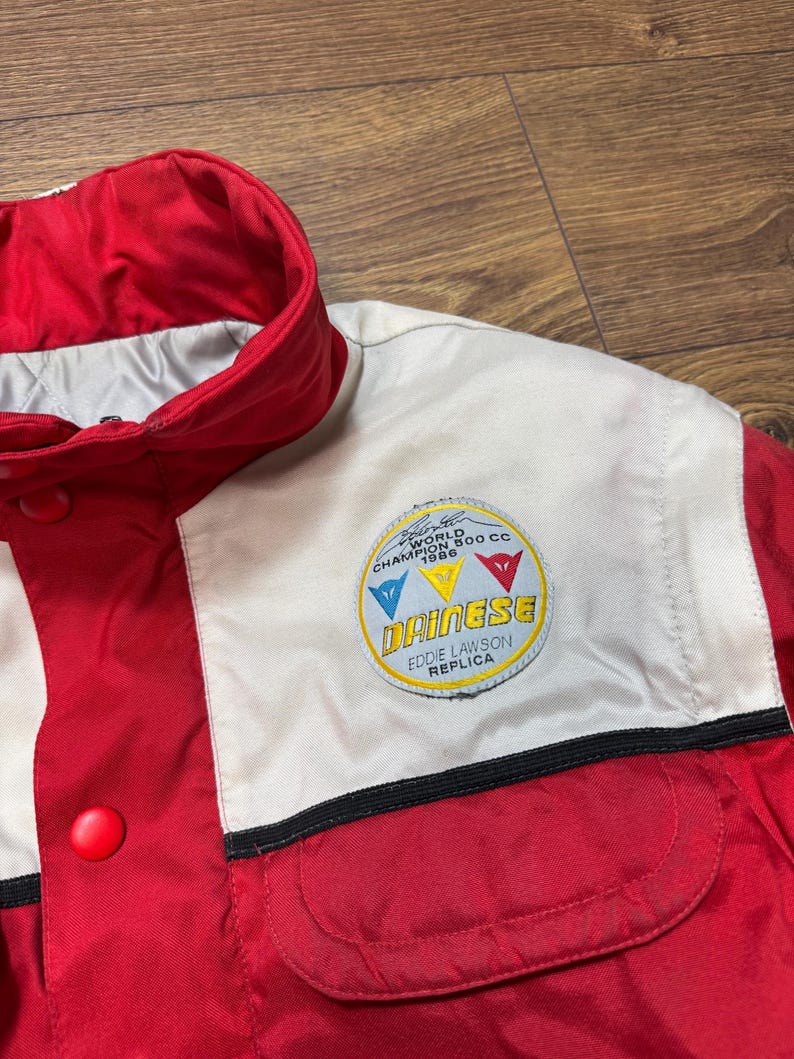 May include: Red and white racing jacket with a button closure and a small pocket. The jacket features a patch with the text "Dainese Eddie Lawson Replica" and a logo with the text "World Champion 500 CC 1986".