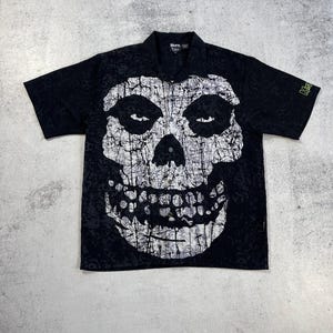 Peut inclure: Chemise noire à manches courtes ornée d'un grand motif de crâne blanc. La chemise a un col et des boutons sur le devant. Le logo Misfits est visible sur la manche.