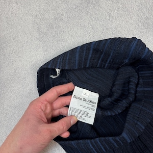 Puede incluir: Prenda de punto acanalado azul marino oscuro con sutiles rayas azules. La prenda tiene una etiqueta que dice "Acne Studios". La tela parece ser una mezcla de materiales. La prenda est&aacute; doblada y sostenida por una mano.
