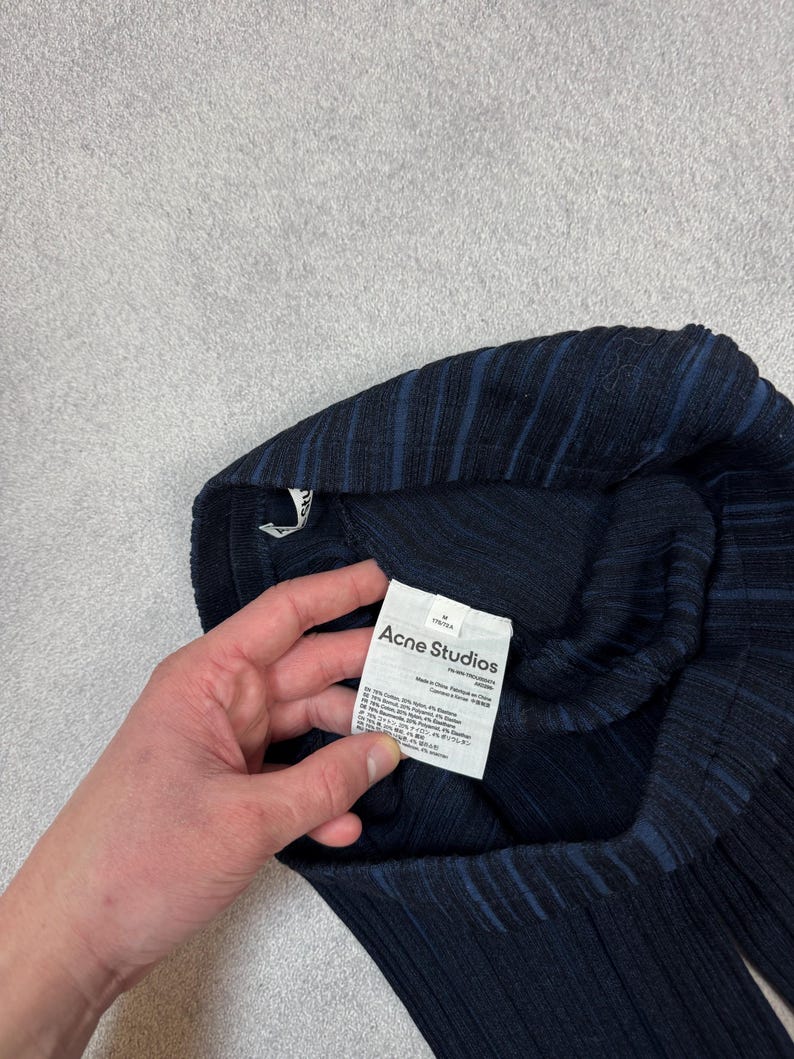 Puede incluir: Prenda de punto acanalado azul marino oscuro con sutiles rayas azules. La prenda tiene una etiqueta que dice "Acne Studios". La tela parece ser una mezcla de materiales. La prenda est&aacute; doblada y sostenida por una mano.