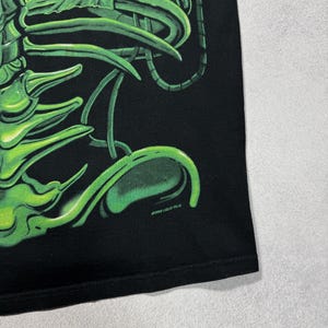 Puede incluir: Camiseta negra con un dise&ntilde;o de esqueleto estilizado en verde ne&oacute;n. El gr&aacute;fico tiene una apariencia brillante y tridimensional. El texto "&copy;1998 LIQUID BLUE" est&aacute; impreso en blanco cerca del dobladillo inferior. La camiseta est&aacute; hecha de un material suave.