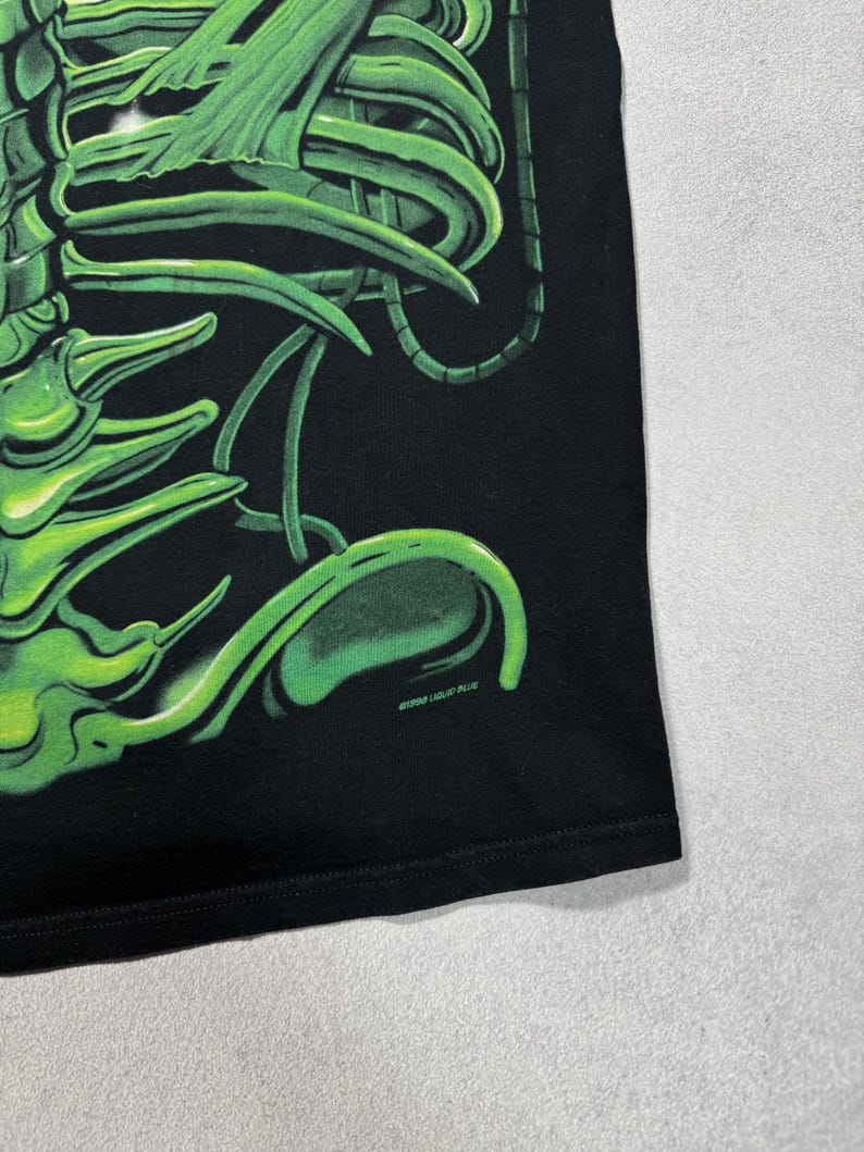 Puede incluir: Camiseta negra con un dise&ntilde;o de esqueleto estilizado en verde ne&oacute;n. El gr&aacute;fico tiene una apariencia brillante y tridimensional. El texto "&copy;1998 LIQUID BLUE" est&aacute; impreso en blanco cerca del dobladillo inferior. La camiseta est&aacute; hecha de un material suave.
