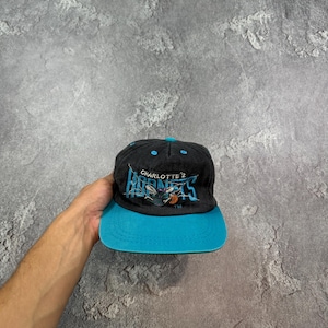 Op de afbeelding: Een zwarte en blauwgroene Charlotte Hornets baseball cap. De cap heeft het logo van het team en de tekst "CHARLOTTE HORNETS" in blauwgroen en wit. De klep is blauwgroen en de cap is gemaakt van een duurzame stof.