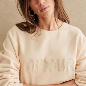 May include: A cream-colored long-sleeve sweatshirt featuring the word AMOUR embossed in a tonal, raised font across the chest. The garment has a classic crew neckline and raglan sleeves, providing a clean, uniform look in a neutral shade.