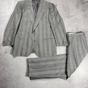 May include: A light gray, two-piece suit with a subtle pinstripe pattern. The jacket has a notched lapel and two buttons. The trousers are pleated. The suit is laid out on a light gray surface.