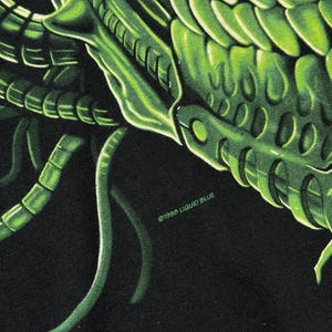 Puede incluir: Primer plano de una camiseta negra con una vibrante ilustraci&oacute;n verde detallada del exoesqueleto de una criatura. El dise&ntilde;o incluye patrones y texturas intrincados, con el texto "&copy;1996 LIQUID BLUE".