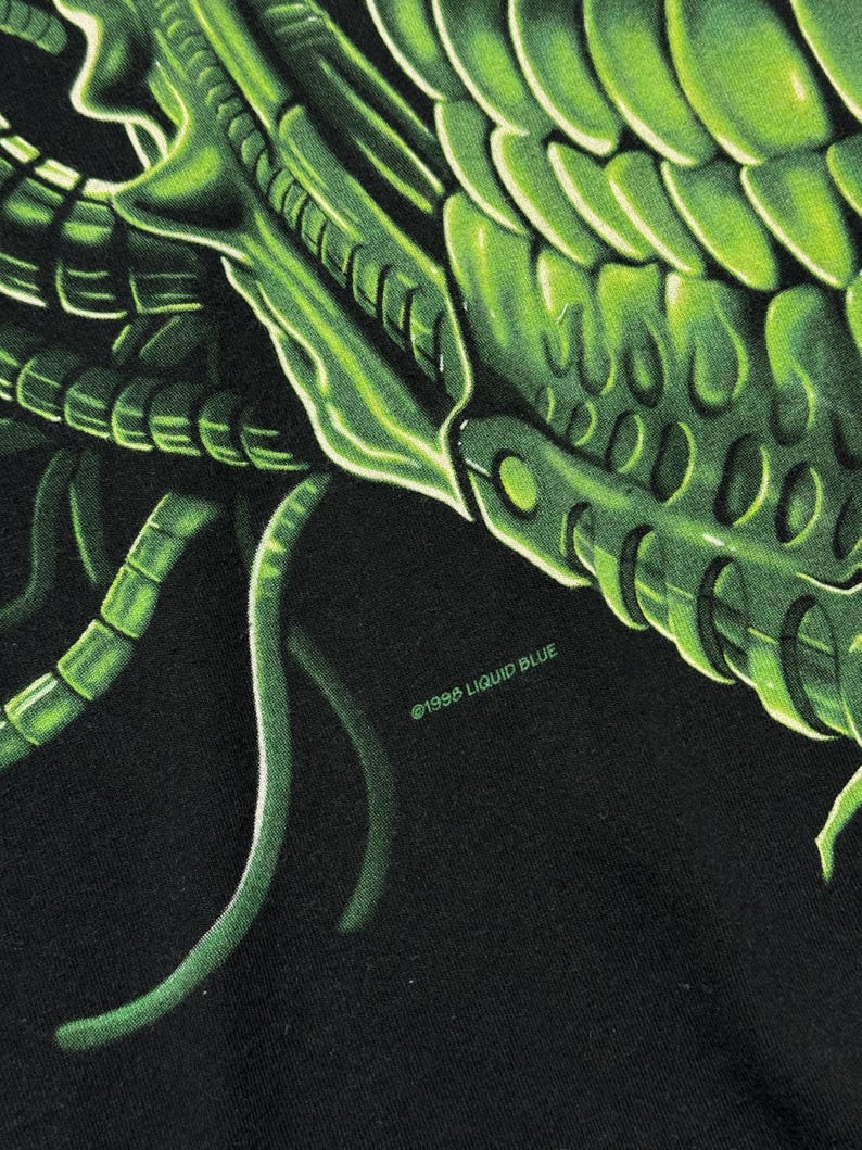Puede incluir: Primer plano de una camiseta negra con una vibrante ilustraci&oacute;n verde detallada del exoesqueleto de una criatura. El dise&ntilde;o incluye patrones y texturas intrincados, con el texto "&copy;1996 LIQUID BLUE".