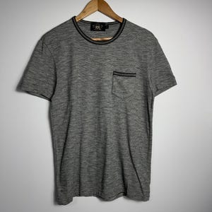 May include: A grey short-sleeved t-shirt with a crew neck and a pocket on the chest. The shirt is hanging on a wooden hanger. The trim around the neck and pocket is black.