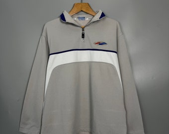 Può includere: Una maglia grigia a maniche lunghe con colletto con zip. La maglia presenta una striscia orizzontale bianca sul petto, una linea di accento blu navy e un piccolo logo. Il colletto e i polsini hanno finiture blu navy.