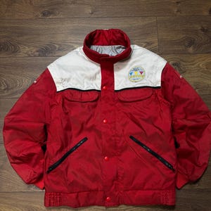 May include: A red and white vintage jacket with a stand-up collar. The jacket has a white upper chest and collar, with a logo patch. It features black zippered pockets and red snap closures. The jacket is made of a shiny material.
