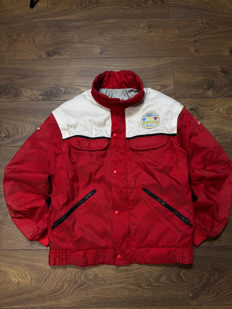 May include: A red and white vintage jacket with a stand-up collar. The jacket has a white upper chest and collar, with a logo patch. It features black zippered pockets and red snap closures. The jacket is made of a shiny material.