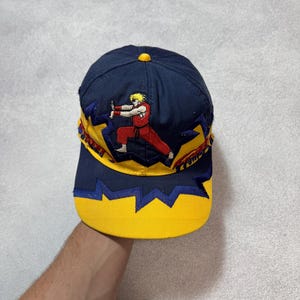 Puede incluir: Gorra de béisbol azul marino con visera y detalles amarillos. La gorra presenta una imagen bordada de un personaje en posición de combate, con pelo amarillo y un atuendo rojo. El diseño incluye estrellas amarillas y formas de rayo azules.