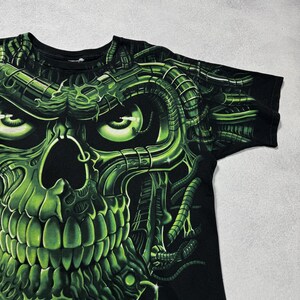 Puede incluir: Camiseta negra con un dise&ntilde;o de calavera verde ne&oacute;n. La calavera tiene una est&eacute;tica mec&aacute;nica y futurista con detalles y sombreados intrincados. El gr&aacute;fico cubre la parte delantera de la camiseta, creando un aspecto audaz y llamativo.