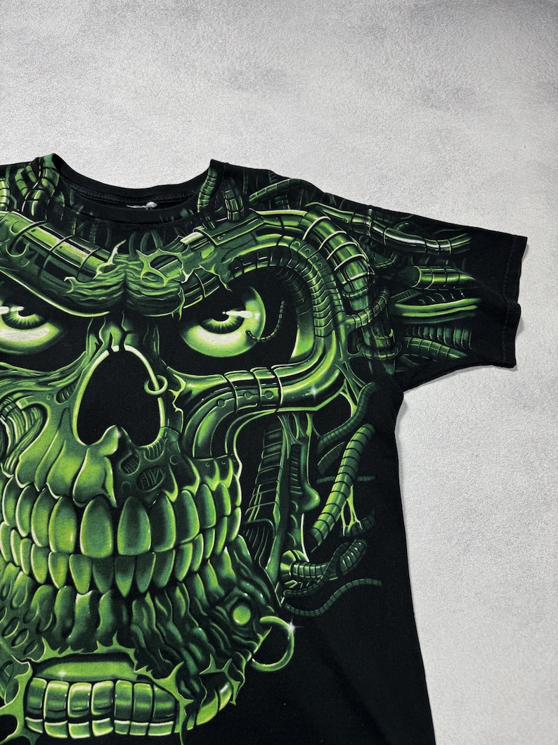 Puede incluir: Camiseta negra con un dise&ntilde;o de calavera verde ne&oacute;n. La calavera tiene una est&eacute;tica mec&aacute;nica y futurista con detalles y sombreados intrincados. El gr&aacute;fico cubre la parte delantera de la camiseta, creando un aspecto audaz y llamativo.