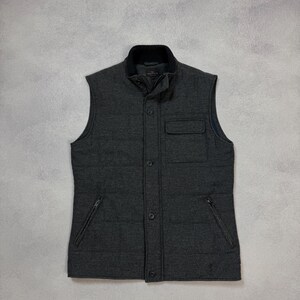 May include: A dark gray quilted vest with a black collar and a button-up front. The vest has two zippered pockets and a chest pocket. The vest is sleeveless and appears to be made of a soft, warm material.